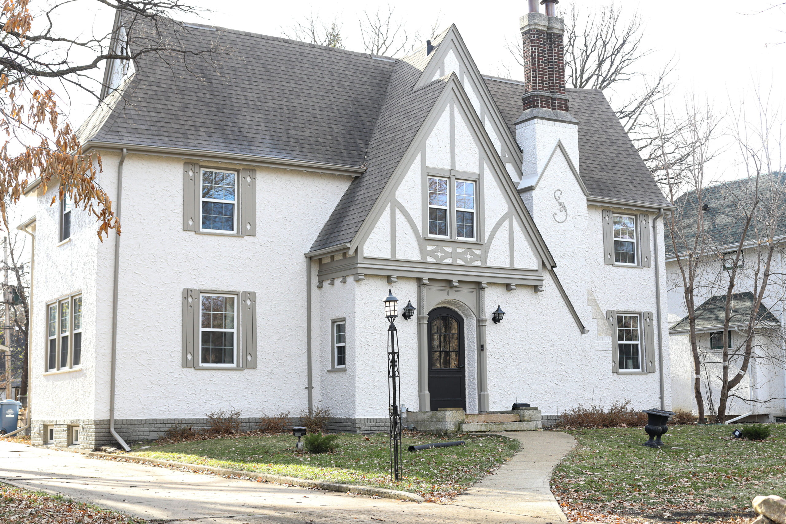 A white stucco Tudor-style house with gray trim, a dark front door, and a chimney, surrounded by bare trees and a grassy yard in autumn.
