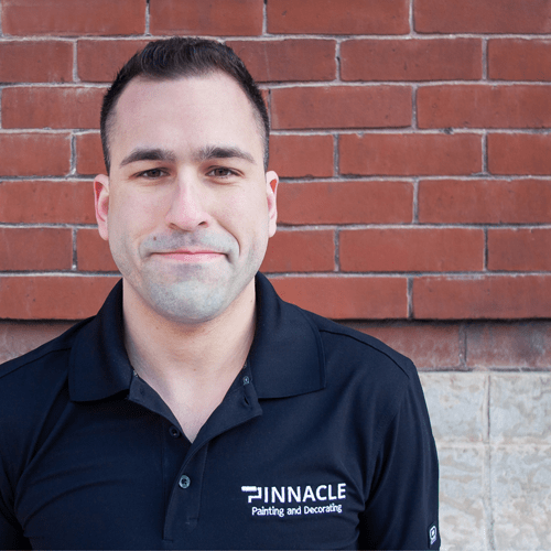 A man in a black polo shirt with 'Pinnacle Painting and Decorating' logo stands against a red brick wall.